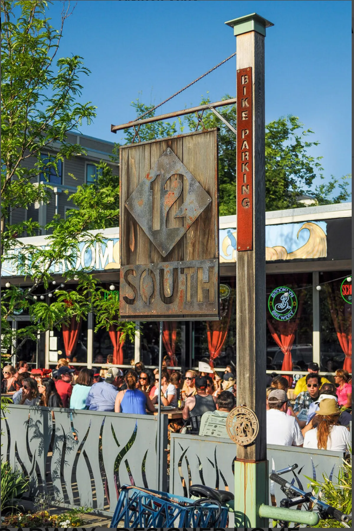 The photo features the lively 12 South neighborhood, with a prominent '12 South' sign, a busy sidewalk, and a crowd of people enjoying outdoor dining. The street scene is packed with energy, showing people biking, socializing, and soaking up the local atmosphere. Bike racks and patio seating reflect the area's walkability and outdoor lifestyle.