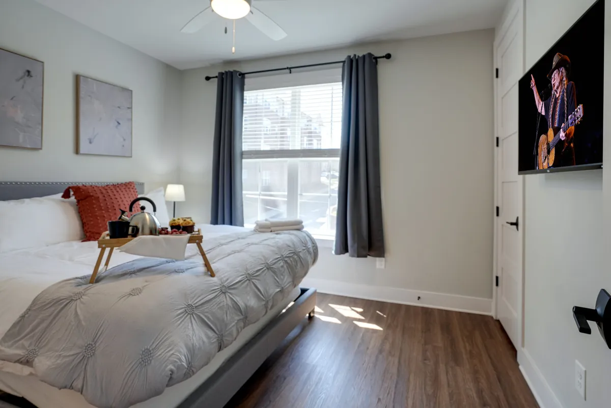 A bright and inviting bedroom featuring a plush bed adorned with decorative pillows, a breakfast tray, and natural light streaming through a window. A TV visible in the scene adds a touch of home comfort, showing a musician performing. The setting is clean, cozy, and welcoming—ideal for restful sleep and relaxation.