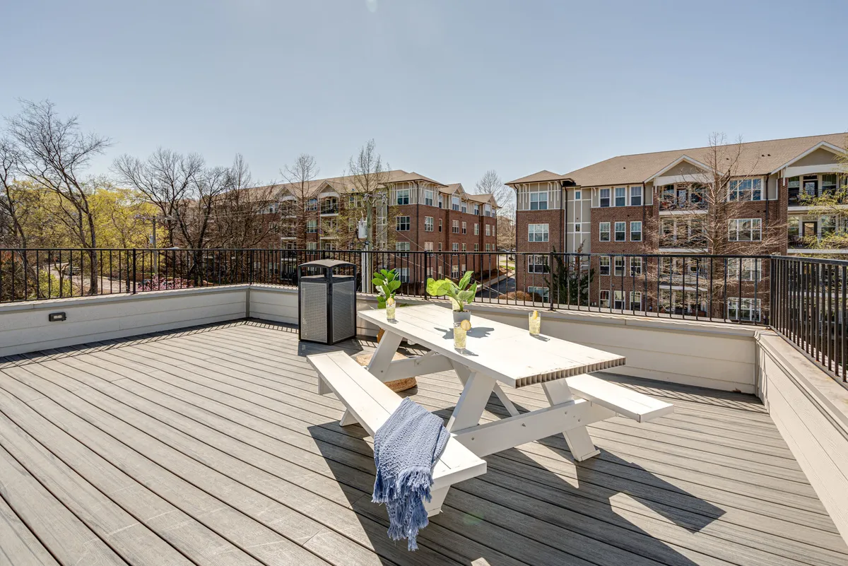 An outdoor dining table is set amidst green plants on a spacious deck, with a casual vibe perfect for group meals or relaxed gatherings. The scene is well-lit and hints at both day and evening use, with city views visible beyond the railing.