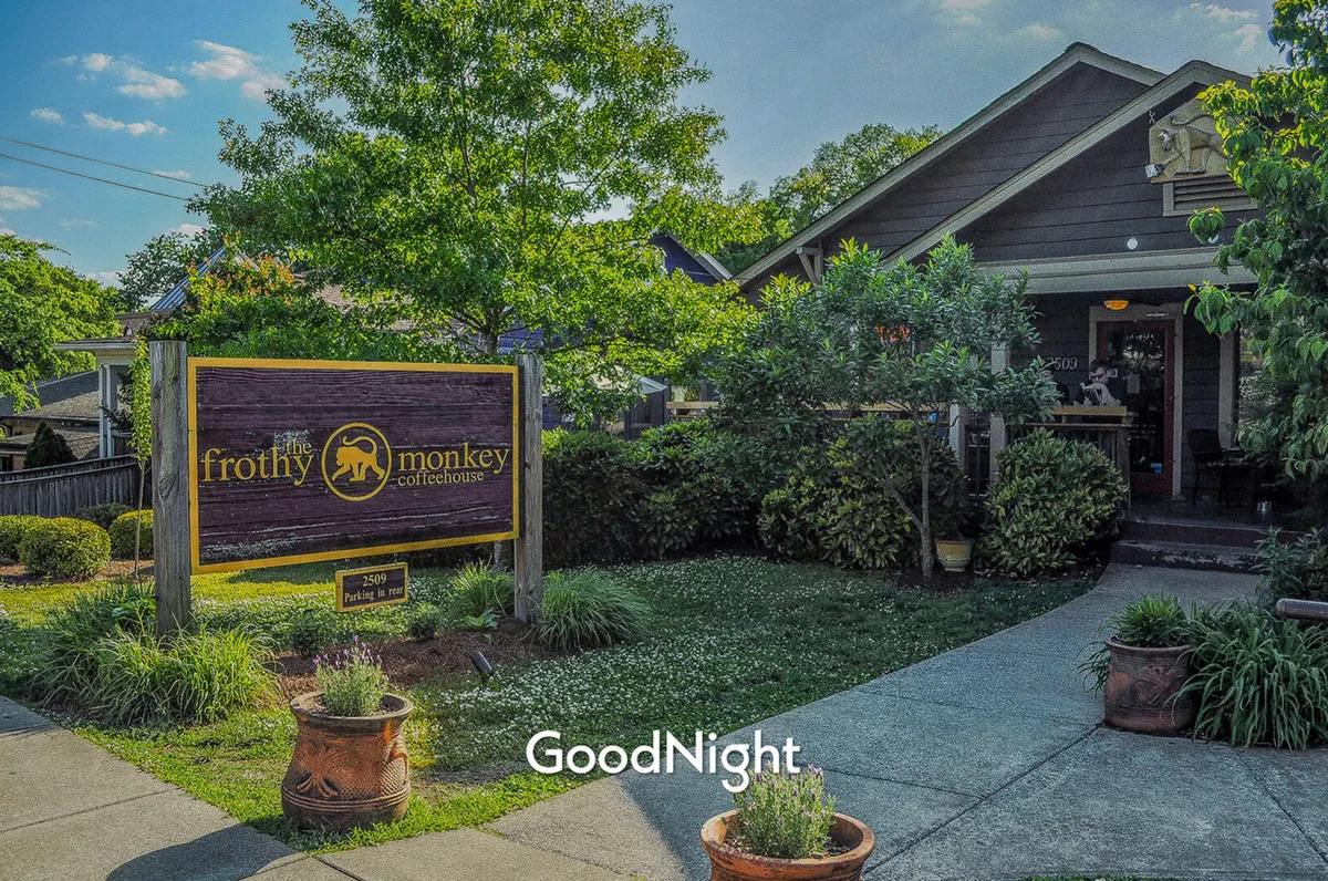 A cozy, welcoming coffeehouse entrance surrounded by lush greenery and decorative pots. The signage for 'the frothy monkey coffeehouse' is prominent, suggesting a popular local spot for relaxing, socializing, or working remotely.