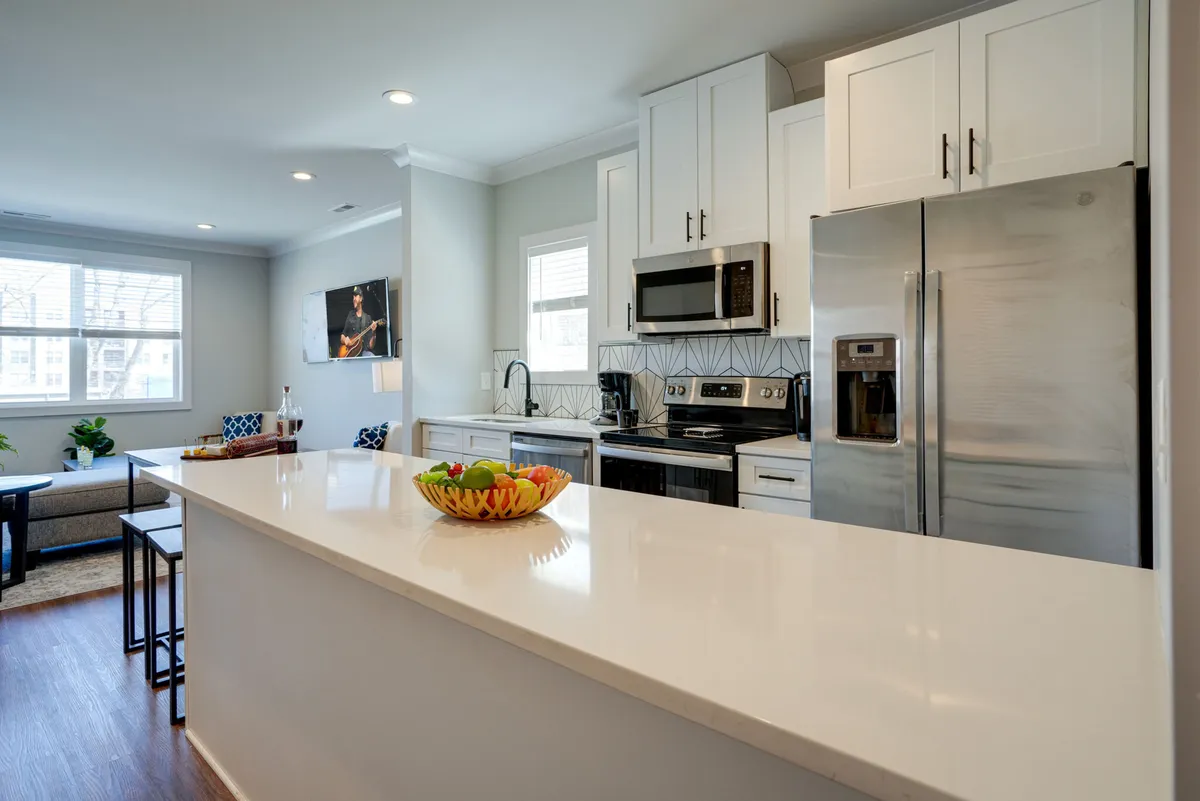An inviting open-concept living space with a kitchen island in the foreground, featuring stainless steel appliances, a fruit bowl, and a visible living area with plush seating. Natural light pours in through windows, creating a warm, cozy atmosphere perfect for socializing or relaxing.