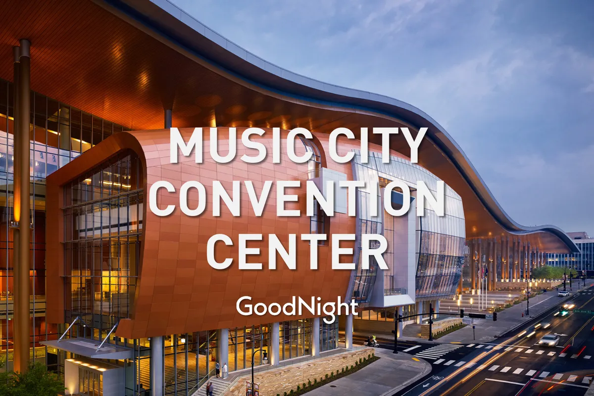 A striking modern building with a dynamic architectural design, featuring large glass windows and a curved roof. The lighting suggests an evening setting, with the city skyline visible in the background. The exterior reflects a space commonly used for conferences, exhibitions, and community events.